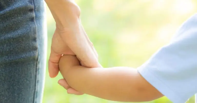 Close up photo of a Mom or nanny holding the hand of a young child, and the photo is zoomed in on their hands. The background is a spring day in Colorado at the park.