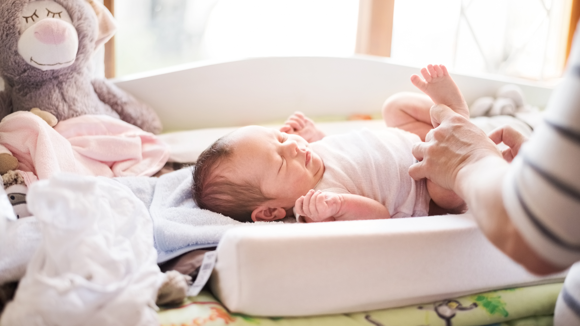 A nanny changing a sleepy baby's diaper on a changing mat surrounded by soft toys