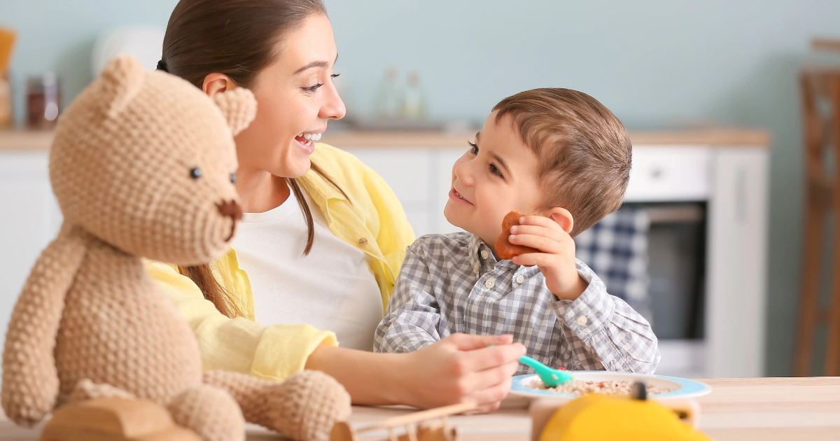 Mother smiling and feeding young child at the table while the child eats and holds a snack next to a teddy bear